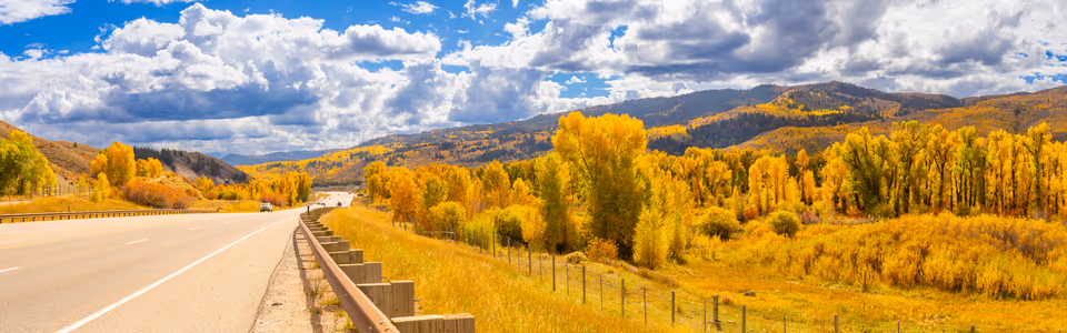 A highway passing through bright golden autumn trees beneath a dramatic mountain sky