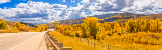 A highway passing through bright golden autumn trees beneath a dramatic mountain sky