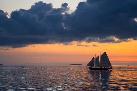 A silhouetted sailboat crossing calm water beneath storm clouds at sunset